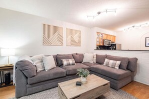 A modern living room with a sectional sofa, a coffee table, and a kitchen area in the background. - Beautiful 2BDR townhouse | Private hot tub | Pets (Whistler)