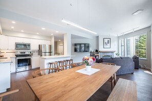 Une cuisine moderne équipée d’appareils électroménagers en acier inoxydable, un coin repas avec des chaises en bois et un canapé gris dans un salon lumineux doté de grandes fenêtres.