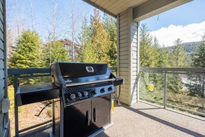 A barbecue grill on a balcony with a view of trees and a house in the background. - Large deck | Private Hot Tub | BBQ | Pet friendly (Whistler)