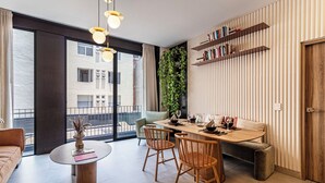 A modern living room with a large window, a dining area with a wooden table and chairs, a sofa, and a bookshelf. - San Luis by Kukun Apartments (Ciudad de México)