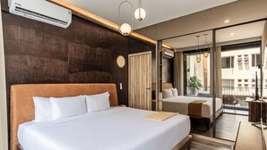 A hotel room with a large bed, a desk, a chair, a window with a view of buildings, and an air conditioning unit. - San Luis by Kukun Apartments (Ciudad de México)
