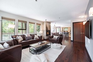 A spacious living room with a fireplace, large windows, and a view of the outdoors. - Large patio | BBQ | Hot tub | Free parking (Whistler)