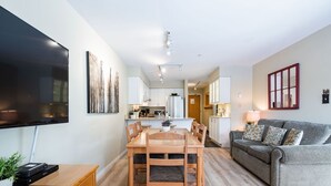 Un soggiorno moderno con TV a schermo piatto, zona pranzo con mobili in legno, un divano grigio e una cucina sullo sfondo.