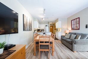 A modern living room with a flat-screen TV, a dining area with wooden furniture, a grey sofa, and a kitchen in the background. - Stunning 2 BDR | Steps to Blackcomb | Free Parking | Shared hot tub (Whistler)