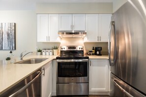 A compact kitchen with white cabinets, a stainless steel refrigerator, a built-in oven, and a coffee maker. - Stunning 2 BDR | Steps to Blackcomb | Free Parking | Shared hot tub (Whistler)