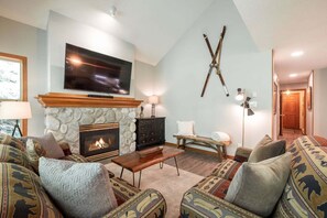 A living room with a stone fireplace, a flat-screen TV, a bench, and patterned sofas. - Close to Blackcomb skiing | Private hot tub | BBQ (Whistler)