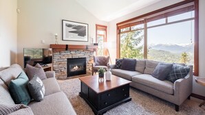 Ein Wohnzimmer mit Kamin aus Stein, einer Couch, einem Couchtisch und einem großen Fenster mit Blick auf die Berge.
