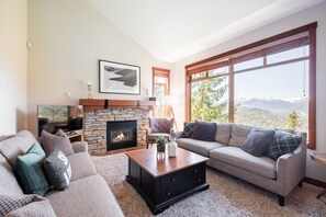 Ein Wohnzimmer mit Kamin aus Stein, einer Couch, einem Couchtisch und einem großen Fenster mit Blick auf die Berge.
