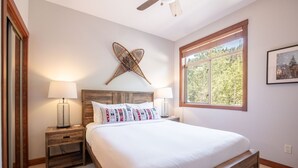 Ein Schlafzimmer mit Holz-Kopfteil, einem großen Fenster mit Blick auf Bäume, zwei Nachttischen mit Lampen und einem Wandkunstwerk.