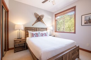 Ein Schlafzimmer mit Holz-Kopfteil, einem großen Fenster mit Blick auf Bäume, zwei Nachttischen mit Lampen und einem Wandkunstwerk.