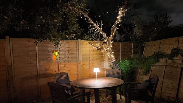 En træterrasse udendørs med et rundt bord og stole, lyskæder og en lanterne.