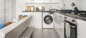 A compact kitchen with a built-in washer and dryer, a stove, and a countertop with various appliances. - Relaxing 5R Flat in Oxford | Near Bus Stop | ST (Oxfordshire)