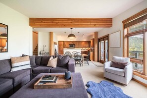 A modern living room with a sofa, a coffee table with books, a dining area with chairs, and a kitchen in the background. - Pets | BBQ | Sundeck | Private garage | Ski-in/out (Whistler)