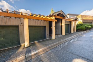 A row of wooden garage doors with a wooden pergola above, a paved driveway, and a clear sky. - Pets | BBQ | Sundeck | Private garage | Ski-in/out (Whistler)