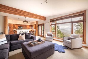 A spacious living area with a sofa, ottoman, and two armchairs, a kitchen with wooden cabinets, and large windows offering a mountain view. - Pets | BBQ | Sundeck | Private garage | Ski-in/out (Whistler)