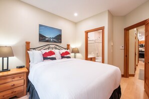 A bedroom with a bed, bedside lamps, a nightstand, a wooden dresser, a painting on the wall, and a bathroom visible through an open door. - Pets | BBQ | AC in bedrooms | Superb location (Whistler)