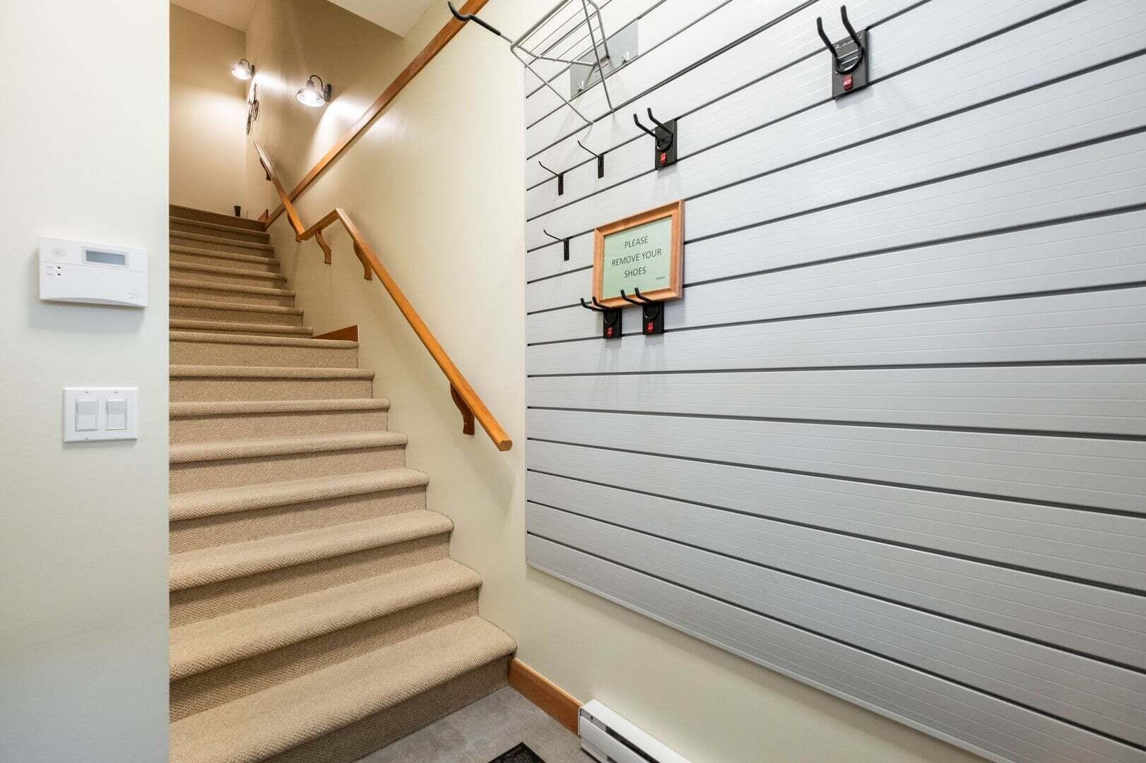 A staircase with a wooden handrail and beige carpeted steps, leading to a room with a grey wall and a sign instructing to remove shoes.
