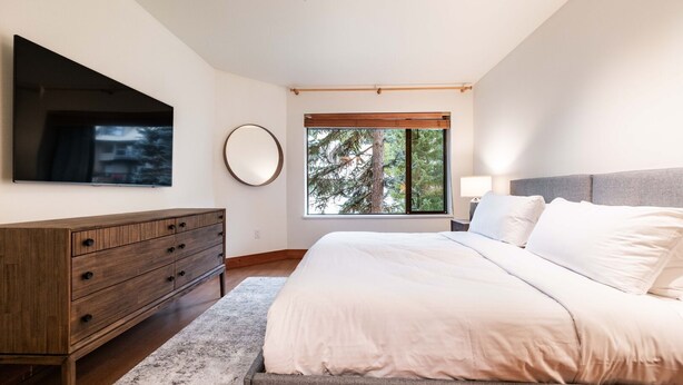 Une chambre à coucher comprenant un lit, une commode, un miroir, un téléviseur et une fenêtre donnant sur des arbres.