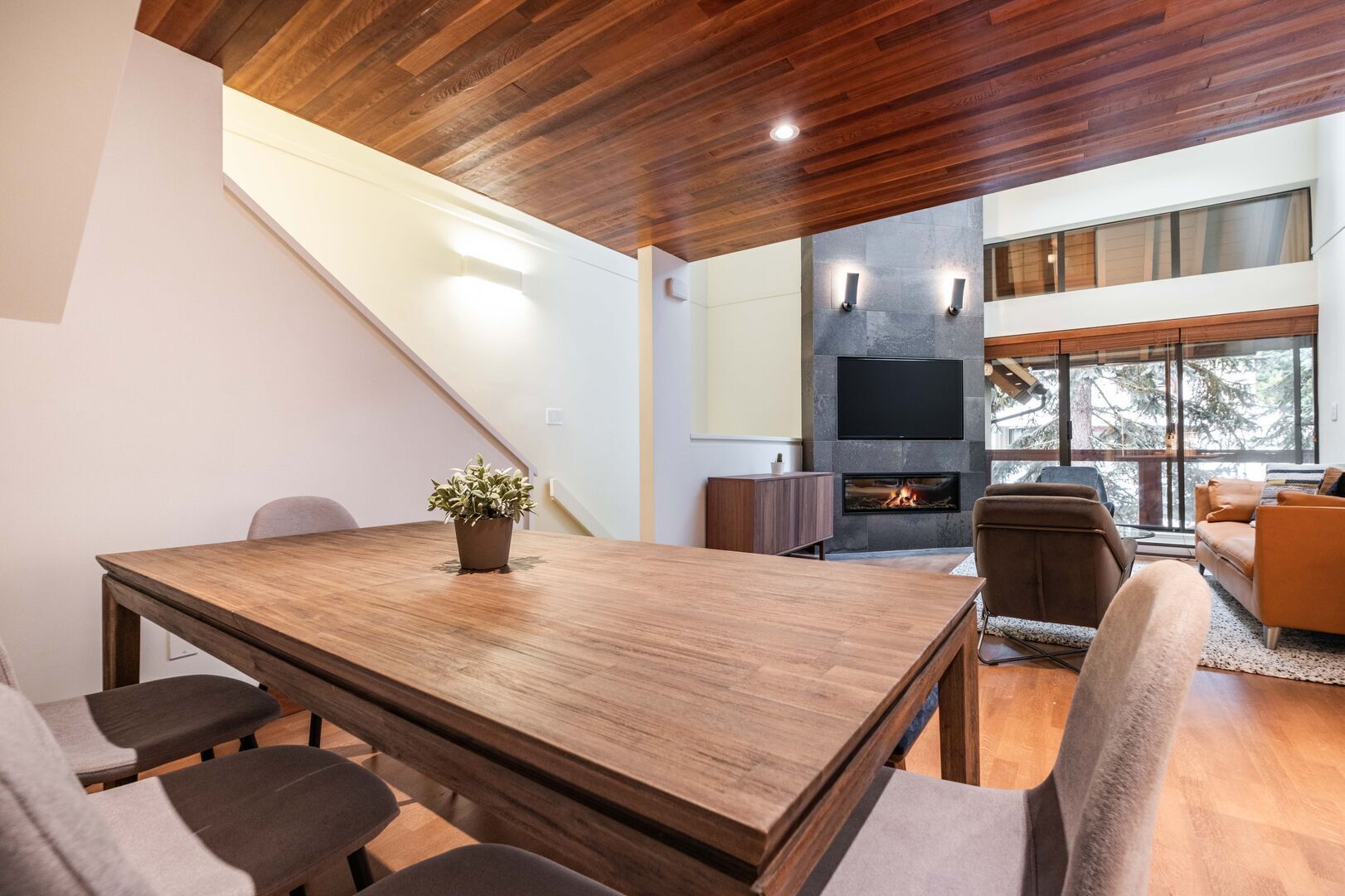 Ein modernes Interieur mit einem Holztisch, einem Kamin und einem großen Fenster mit Blick auf Bäume.