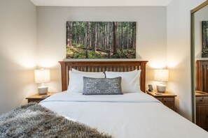 Ein Schlafzimmer mit einem hölzernen Kopfende, einem Bett mit weißer Bettwäsche, zwei Nachttischlampen und einem Waldgemälde über dem Bett.