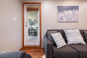 Ein Wohnzimmer mit grauem Sofa, gemusterten Kissen, einem Gemälde einer verschneiten Landschaft und einer Tür zu einem Außenbereich mit Stühlen und einem Tisch.