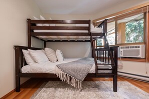 Ein Stockbett mit Holzrahmen, weißer Bettwäsche und einer grauen Tagesdecke. Ein Fenster mit Jalousien und eine Klimaanlage befinden sich an der Wand.