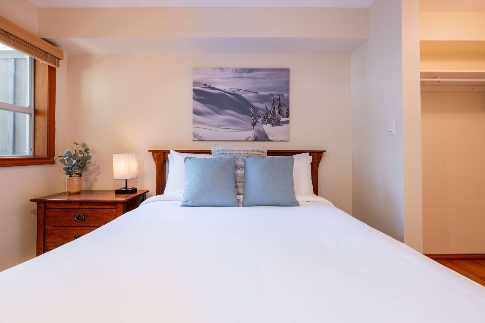 A neatly made bed with a headboard, a bedside table with a lamp, a window with a view, and a painting on the wall.