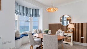 Un comedor con una mesa redonda, sillas tapizadas en gris, un aparador de madera, un espejo y vista al mar a través de una ventana con un valance a rayas.