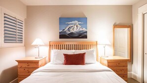 Quarto com cama de madeira, roupa de cama branca, pintura de montanhas na parede e duas mesas de cabeceira com abajures.