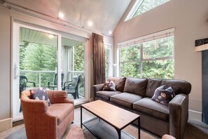 A cozy living room with a sofa, a chair, a coffee table, and large windows offering a view of greenery. - Free Parking | Steps to Blackcomb | Shared Hot Tub (Whistler)