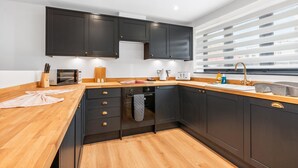 Une cuisine moderne avec des meubles de couleur sombre, des plans de travail en bois, un four encastré et une fenêtre équipée de stores.