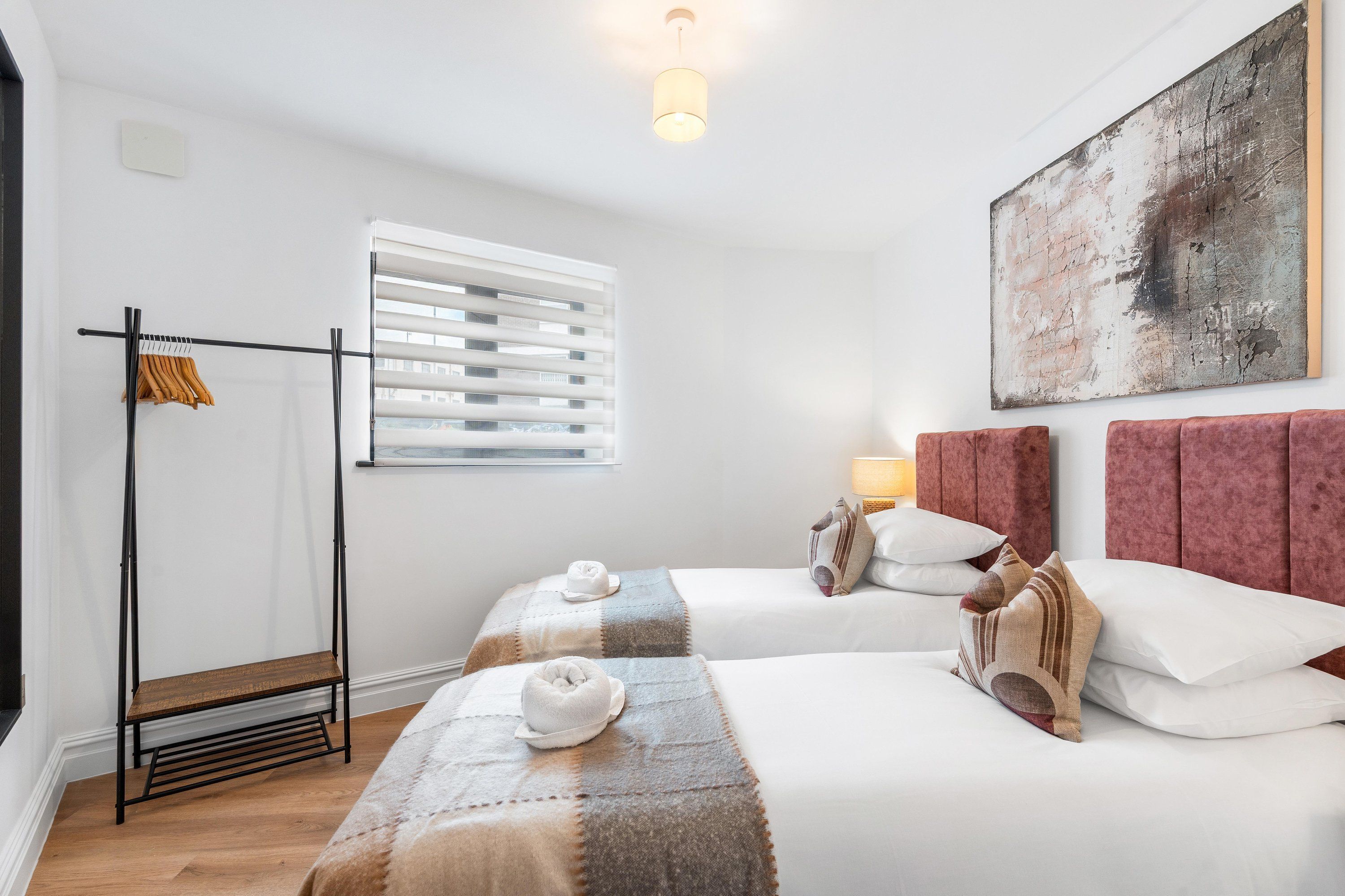 Une chambre d’hôtel avec deux lits, une fenêtre avec des stores, un porte-manteau et un tableau au mur.
