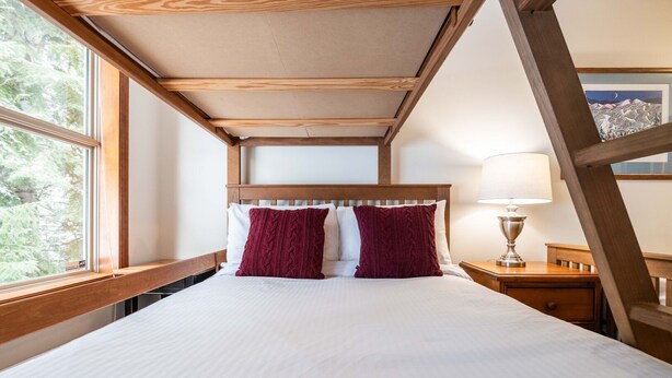 Ein Schlafzimmer mit einem Bett, einem Holzkopfstück, einem Nachttisch mit Lampe und einem Fenster mit Blick auf Bäume.