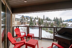 A balcony with red chairs, a grill, and a view of snow-covered houses and trees. - Large deck with BBQ | Hot Tub | Golf Course (Whistler)
