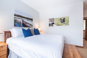 A bedroom with a large bed, a wooden nightstand, a lamp, and two wall-mounted pictures. - Large deck with BBQ | Hot Tub | Golf Course (Whistler)