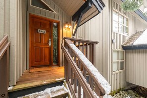 A wooden front door with a small porch, a wooden staircase with snow on the steps, and a window with shutters. - Large deck with BBQ | Hot Tub | Golf Course (Whistler)