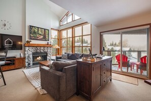 A cozy living room with a stone fireplace, a large window, and a balcony with red chairs. - Large deck with BBQ | Hot Tub | Golf Course (Whistler)