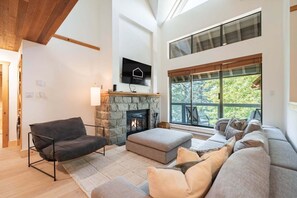 A modern living room with a stone fireplace, a large window with a view of trees, a mounted TV, and a cozy seating area. - BBQ | Free Parking | Spacious | Forest views (Whistler)