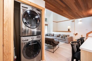 A compact living space with a stacked washer and dryer, a sofa, a dining area with chairs, and a wooden ceiling. - BBQ | Free Parking | Spacious | Forest views (Whistler)