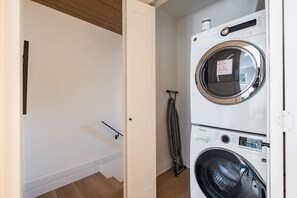 Una sala de lavandería con lavadora y secadora apiladas, un tendedero metálico y una puerta que da a otra habitación.