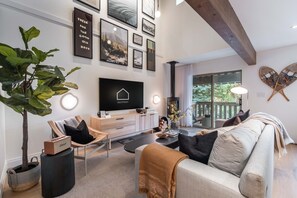 Ein modernes Wohnzimmer mit Flachbildfernseher, bequemen Sitzgelegenheiten, einem großen Fenster und einem Holzbalken an der Decke.