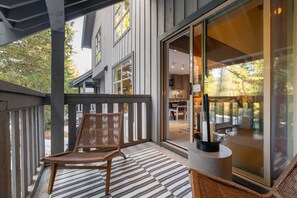 Eine Holzterrasse mit einem Korbstuhl, einer Glastür, die zu einem Raum mit Essbereich führt, und einem gemusterten Teppich.