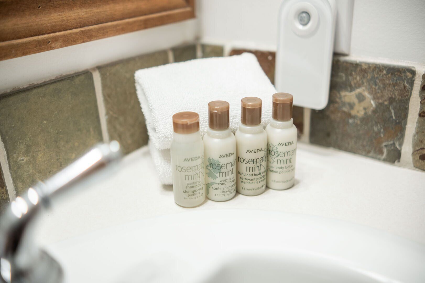 A bathroom sink with three bottles of Aveda body lotion and a white towel.