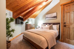 Ein Schlafzimmer mit schrägem Dach, einem Bett mit vielen Kissen, einem Nachttisch, einem an der Wand befestigten Fernseher und einer Holztür.