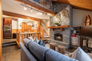 A cozy living room with a stone fireplace, wooden staircase, and a modern kitchen in the background. - Amazing location | Private hot tub | Free parking (Whistler)