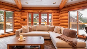 Een gezellig houten blokhutinterieur met een hoekbank, een houten bijzettafeltje en grote ramen met uitzicht op een besneeerd landschap.