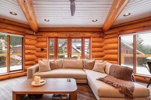 A cozy log cabin interior with a sectional sofa, a wooden coffee table, and large windows offering a view of a snowy landscape. - Luxury log chalet | Private hot tub | Ski-in/out (Whistler)