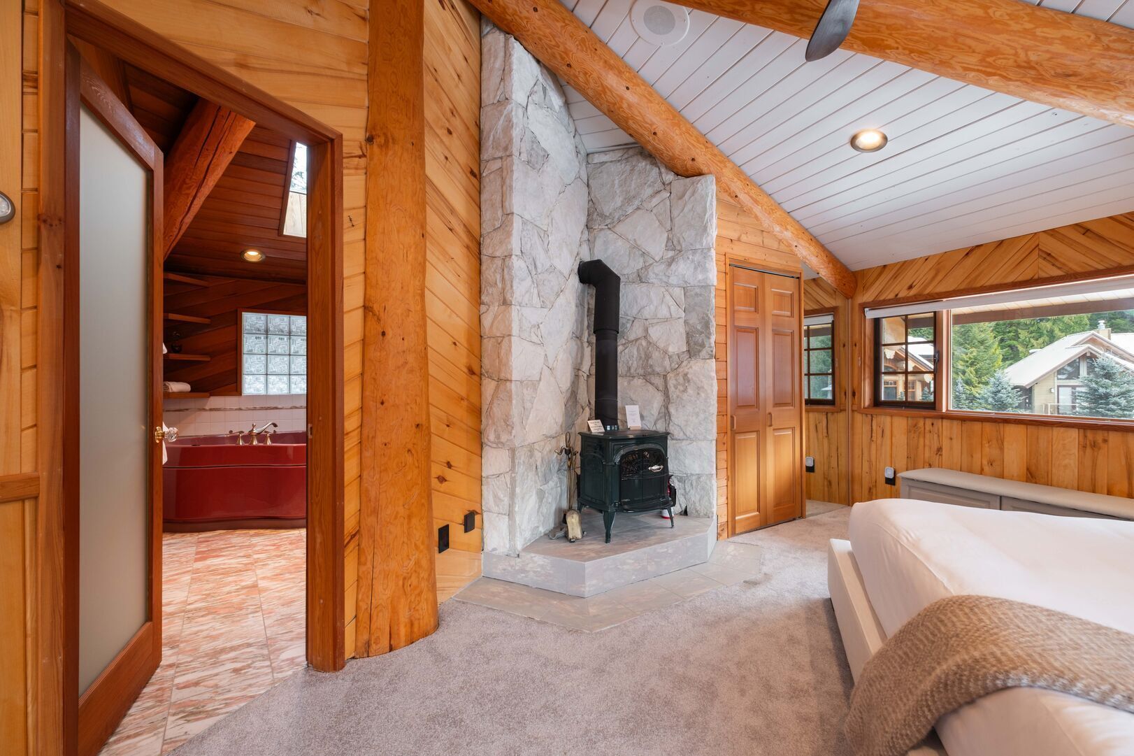 A log cabin interior with a stone fireplace, a red bathtub, and a large window offering a view of the outside.