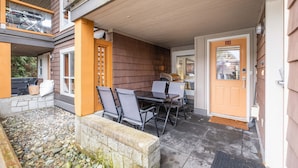 En overdækket udendørs terrasse med et bord, stole, en stenvæg og en trädør med nummer 23.