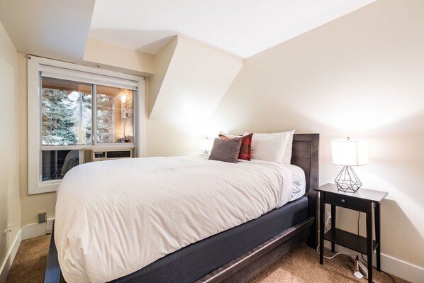 Une chambre avec un grand lit, une fenêtre donnant sur des arbres, une table de chevet avec une lampe et un sol recouvert d’une moquette.